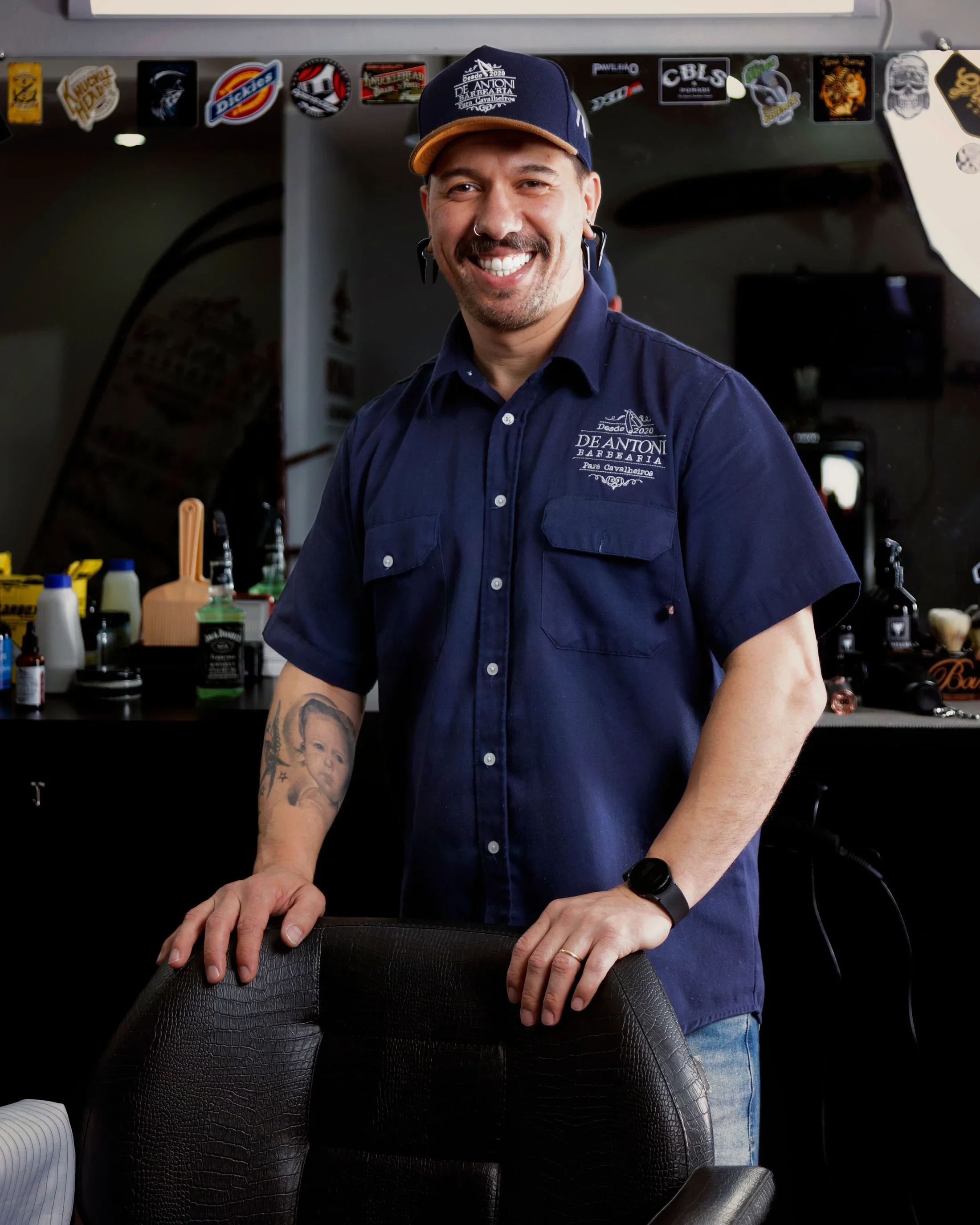 A smiling barber in a blue shirt standing behind a leather barber chair